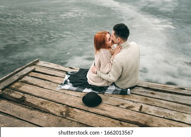 Young loving couple sitting on the pier near a frozen lake. A woman hugs a guy, kissing, in love. Multicultural couple, Asian and European red hair.