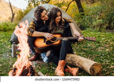 Young loving couple of tourists relaxing near the bonfire in the nature. Handsome man teaches his girl to play guitar on a hike by the campfire. - Powered by Shutterstock