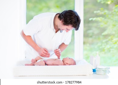 Young Loving Father Changing Diaper Of His Newborn Baby Son, Holding Lotion Bottle In His Hand, Standing In A Sunny Bedroom With A Big Garden View Window 