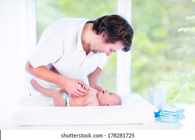 Young Loving Father Changing Diaper Of His Newborn Baby Son, Holding Lotion Bottle In His Hand, Standing In A Sunny Bedroom With A Big Garden View Window 