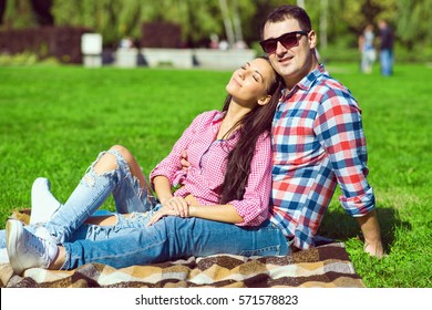 Young loving happy couple in checked shirts, jeans and white sneakers sitting on the green lawn. Smiling girl with closed eyes leaning on her boyfriend. Outdoor shot - Powered by Shutterstock