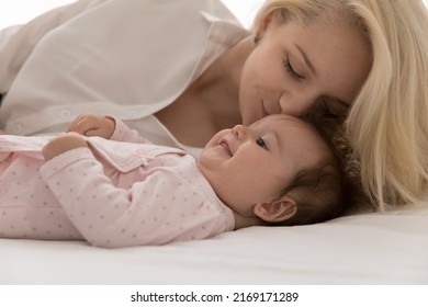 Young Loving Mother Kisses On Forehead Her Lovely Babygirl, Lying Together On Bed, Close Up Cropped Shot. Woman Feeling Pure Love To Newborn Baby, Express Caress. Happy Motherhood, Family Bond Concept