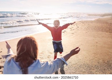 Young Loving Mother Stretched Out Arms To Side Of Smiling Daughter Running Towards Her On Sunny Beach, Happy Mom Ready To Hug Little Girl, Enjoying Spending Time Together On Summer Seaside. Rear View