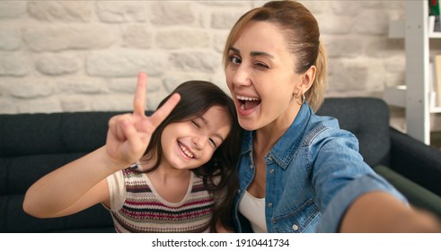 Young Loving Mother Taking Selfie By Phone With Daughter, Making Victory Sign, Sitting On Soft Comfortable Sofa At Home. Woman And Girl Taking Photo Or Video On Smart Phone Camera Smiling Together.