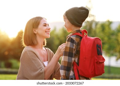 Young Loving Smiling Mother Leading Son Schoolboy With Backpack To First Grade, Mom Sitting Down Next To Her Little Child And Saying Good Bye Before School While Standing Together Outdoor On Sunny Day