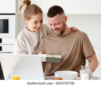 Young loving wife making surprise for husband for anniversary or birthday, congratulating him and giving present while having healthy breakfast in morning at kitchen, happy man opening gift box
