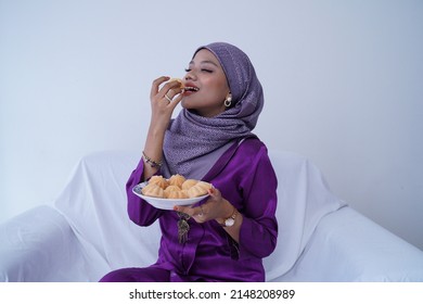 A Young Malay Lady With A Hijab Celebrates Eid Mubarak, Dressed In A Purple Kebaya Dress, Holding Traditional Bahulu Cake, Sitting On A White Sofa White Background.