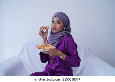 A Young Malay Lady With A Hijab Celebrates Eid Mubarak, Dressed In A Purple Kebaya Dress, Holding Traditional Bahulu Cake, Sitting On A White Sofa White Background.