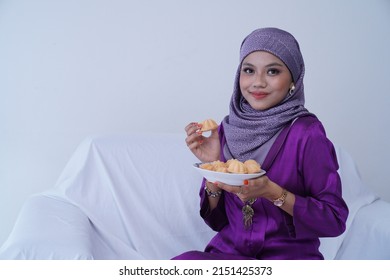 A Young Malay Lady With A Hijab Celebrates Eid Mubarak, Dressed In A Purple Kebaya Dress, Holding Traditional Bahulu Cake, Sitting On A White Sofa White Background.