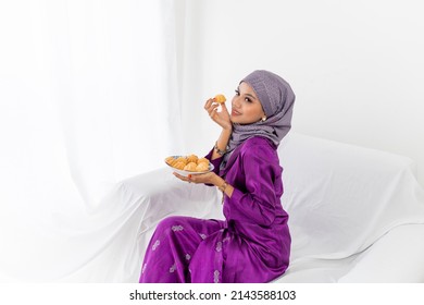 A Young Malay Lady With Hijab Head Covering, Celebrates End Of Ramadan, Dressed In Purple Kebaya Dress, Holding Traditional Bahulu Cake, Sitting On White Sofa, White Background