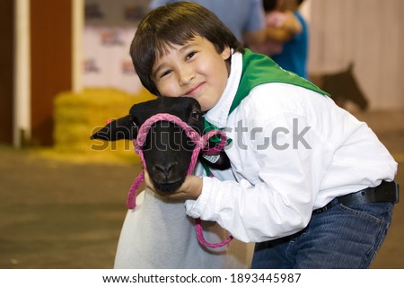 A young male 4-H agricultural club showman hugs his lamb while looking at the camera.