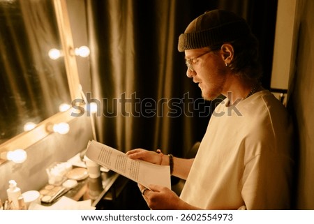 Young male actor reading script in dressing room backstage before performance under warm light next to mirror lined with bulbs and various makeup products