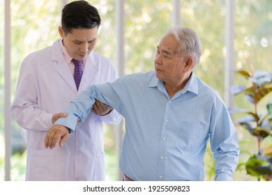 A Young Male Asian Black Hair Doctor Wearing White Lab Coat Holding The Patient Arm And Checking His Elbow When The Senior White Hair Patient Wearing Light Blue Shirt Stand Next To Him .