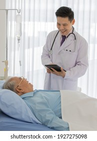 A Young Male Asian Doctor Wearing White Lab Coat With Stethoscope Holding Black Tablet In His Hands Stand Smiling In Front Of Old Fat Patient Lying Down In Hospital Room.