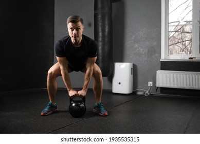 A young male athlete lifts a 20 kg metal kettlebell with two hands from a bent knee position. Training in the gym to raise the metal. Place for your text.