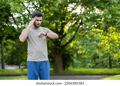 Young male athlete outdoors checking smartwatch. Man appears tired after training session while standing in sunlit park. Concept of fitness, exercise, wearable technology. - Powered by Shutterstock