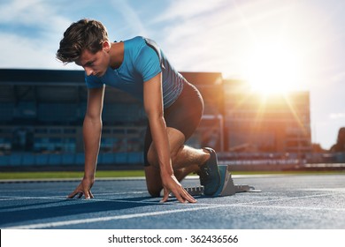 Young Male Athlete At Starting Block On Running Track. Young Man In Starting Position For Running On Sports Track. Sprinter About To Start A Race At Stadium With Sun Flare.