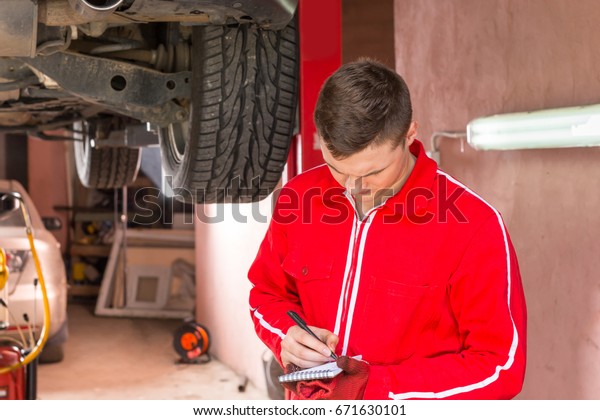 Young male auto mechanic standing making notes\
underneath a lifted car near working equipment for repair and\
diagnostic in auto repair\
service