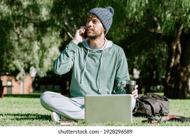 Young Male Backpacker Working Freelance. A Handsome Male With Beanie Calling With His Mobile Phone While Drinking A Coffee. Concept: Digital Nomad Life