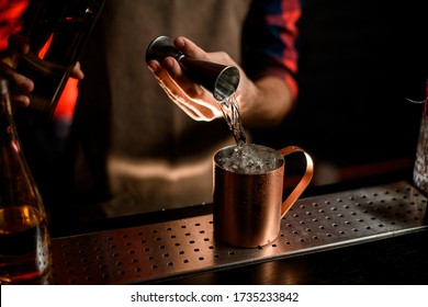 young male bartender in brown apron gently pours drink from metal jigger into stainless cup with ice.