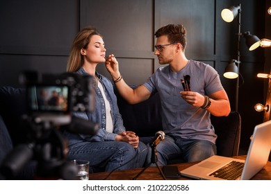 Young Male Beauty Blogger Sitting On Sofa In Front Of A Camera Is Applying Makeup To Beautiful Young Woman