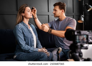 Young Male Beauty Blogger Sitting On Sofa In Front Of A Camera Is Applying Makeup To Beautiful Young Woman