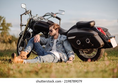 Young Male Biker In Sunglasses Is Sitting On The Groung With A Motor Bike