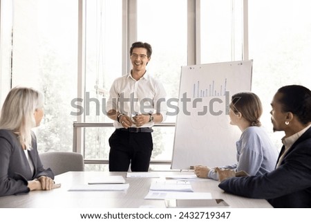 Young male boss make flip chart presentation for company client. Group of multiethnic businesspeople gathered together in office board room take part in seminar workshop led by millennial male trainer 商業照片 © 