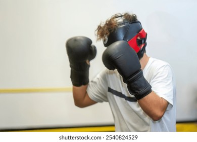 Young male boxer wearing protective headgear and boxing gloves practicing punches during training session in gym, showcasing dedication to boxing and fitness - Powered by Shutterstock