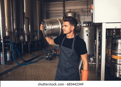 Young Male Brewer Carrying Keg At Brewery. Manual Worker With Metal Beer Barrel Craft Beer Manufacturing Plant.