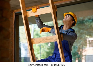 Young Male Builder In Overalls And Hard Hat Looking Focused, Climbing Up The Ladder While Working At Cottage Construction Site