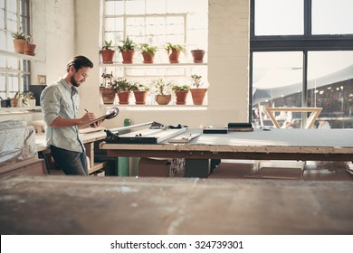 Young Male Business Owner Standing In His Studio Workshop Checking Figures And Stock On His Clipboard On A Sunny Afternoon