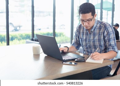 Young Male Businessman Writing On A Notepad In Front Of A Laptop On Wood Desk In Coffee Cafe Shop. Student Using Notebook At College.