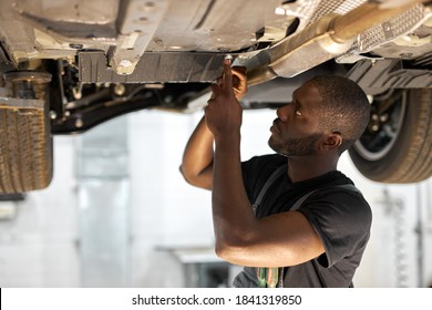 Young Male Car Mechanic In Uniform Checking Car In Automobile Service With Lifted Vehicle, Handsome Hardworking Guy Working Under Car Condition On Lifter. Automotive Car Repair Concept