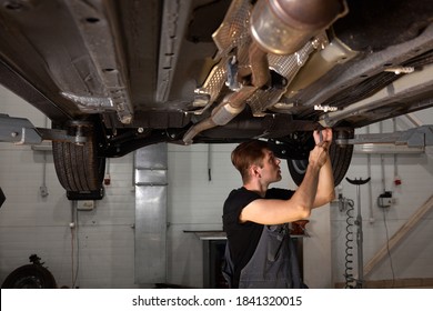 Young Male Car Mechanic In Uniform Checking Car In Automobile Service With Lifted Vehicle, Handsome Hardworking Guy Working Under Car Condition On Lifter. Automotive Car Repair Concept