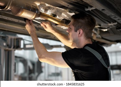 Young Male Car Mechanic In Uniform Checking Car In Automobile Service With Lifted Vehicle, Handsome Hardworking Guy Working Under Car Condition On Lifter. Automotive Car Repair Concept