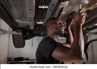 Young Male Car Mechanic In Uniform Checking Car In Automobile Service With Lifted Vehicle, Handsome Hardworking Guy Working Under Car Condition On Lifter. Automotive Car Repair Concept