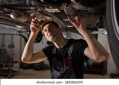 Young Male Car Mechanic In Uniform Checking Car In Automobile Service With Lifted Vehicle, Handsome Hardworking Guy Working Under Car Condition On Lifter. Automotive Car Repair Concept