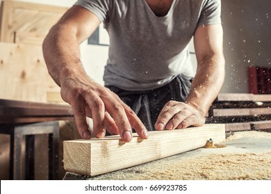 A young male carpenter builder in a gray T-shirt and working overall equals a wooden bar with a milling machine in the workshop, in the background wooden boards