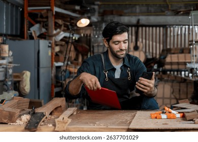 Young male carpenter using smartphone and holding clipboard in woodworking workshop. Concept of modern craftsmanship, multitasking, small business owner, digital tools, and work management