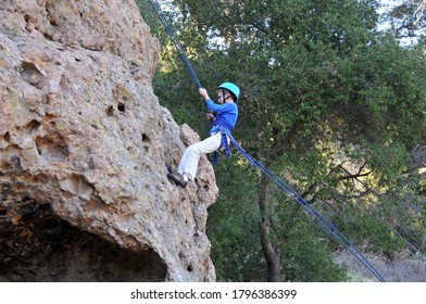 Young Male Caucasian Child Wearing Tan Pants And Blue Long Sleeved Shirt With Blue Helmet, Attached To Safety Ropes Learning To Rock Climb. Fun Healthy Outdoor Activity