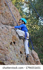 Young Male Caucasian Child Wearing Tan Pants And Blue Long Sleeved Shirt With Blue Helmet, Attached To Safety Ropes Learning To Rock Climb. Fun Healthy Outdoor Activity