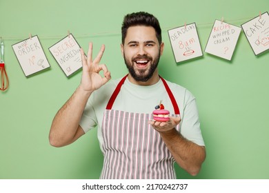 Young male chef confectioner baker man in striped apron hold pink little cake muffin macaroon show ok okay gesture isolated on plain pastel light green background studio portrait. Cooking food concept