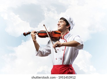 Young male chef playing violin on blue sky background. Handsome chef in white hat and red apron standing with music instrument. Creative culinary performance. High cooking skills concept