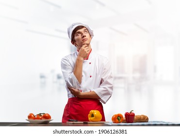 Young Male Chef Standing With Folded Arms And Looking Up Dreamily. Pensive Culinary School Student In White Hat And Red Apron In Light Kitchen Interior. Professional Kitchen Staff Recruitment