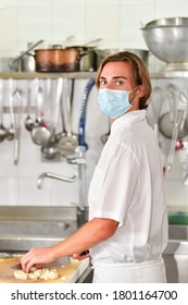 Young Male Chef With A Surgical Mask Using A Knife To Cut Potatoes On A Cutting Board While Looking At The Camera. Cooking And Safety Concept.