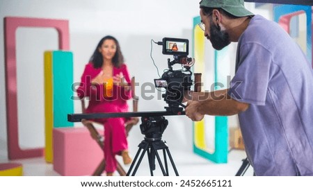 Young Male Cinematographer With Beard, Wearing Cap, Records Vibrant Female Host In Pink Suit During Lively Talk Show Set With Colorful Geometric Shapes, Enhancing Modern Video Production, Storytelling