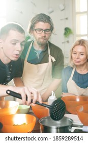 Young Male Cooking Coach And His Two Trainees In Aprons Bending Over Workplace With Electric Stove While Checking If Ingredients Are Ready