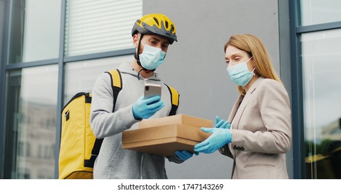 Young Male Courier In Medical Mask Bringing Carton Boxes To Woman On Street And Tapping On Phone To Check Parcel. Food Delivery Service Concept