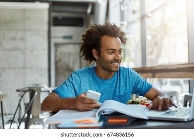 Young male with dark skin and curly hair surrounded with books holding telephone in his hand looking in laptop with smile being happy to find what he needs for project. People, youth, education